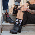 Cropped detail of the seated woman's hands resting on her knee next to her partner's boot on the step, showcasing Dia De Los Muertos Skull socks in Black with pink, blue, and yellow floral skull illustrations. [lf]