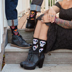 Cropped detail of the seated woman's hands resting on her knee next to her partner's boot on the step, showcasing Dia De Los Muertos Skull socks in Black with pink, blue, and yellow floral skull illustrations. [lf]