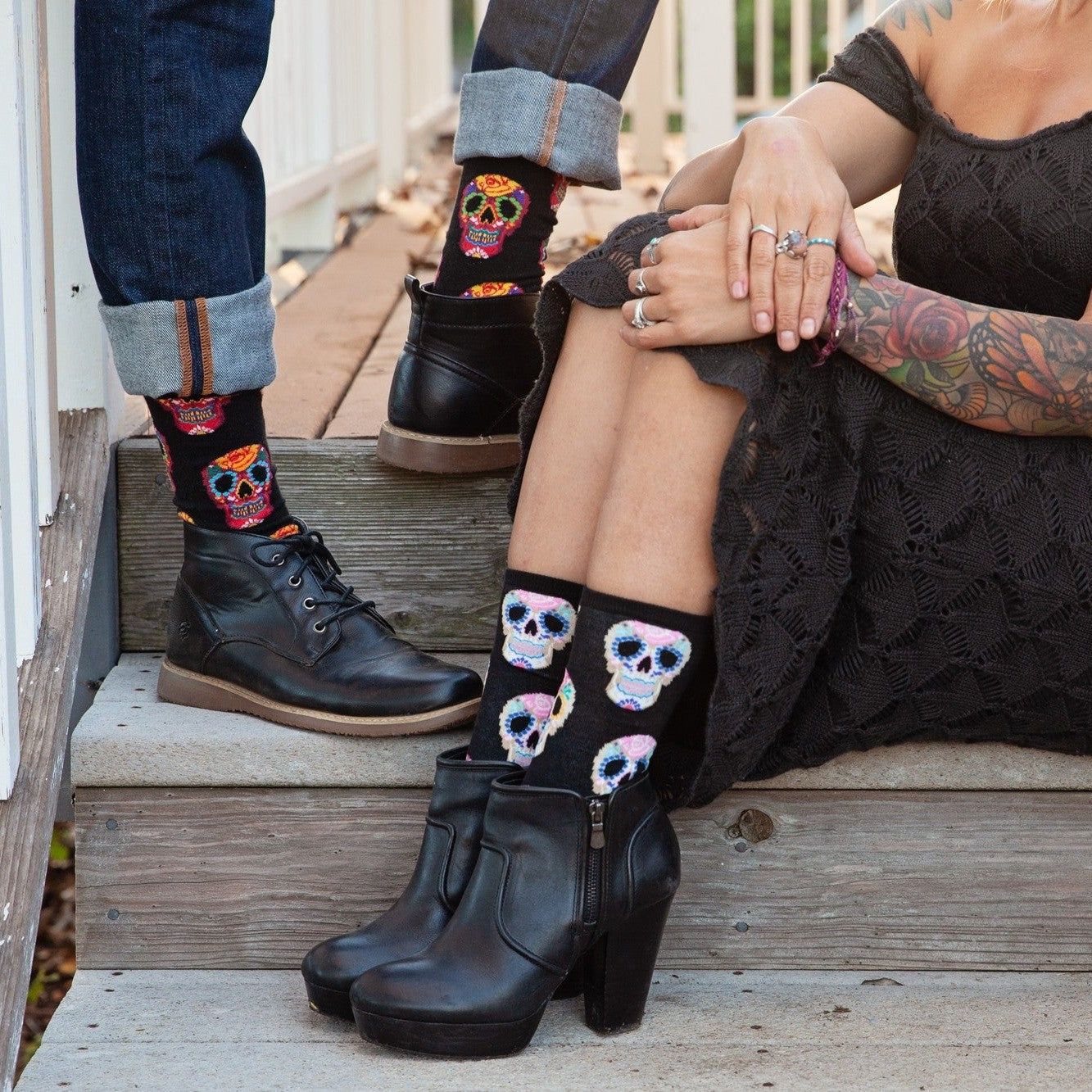 Cropped detail of the seated woman's hands resting on her knee next to her partner's boot on the step, showcasing Dia De Los Muertos Skull socks in Black with pink, blue, and yellow floral skull illustrations. [lf]