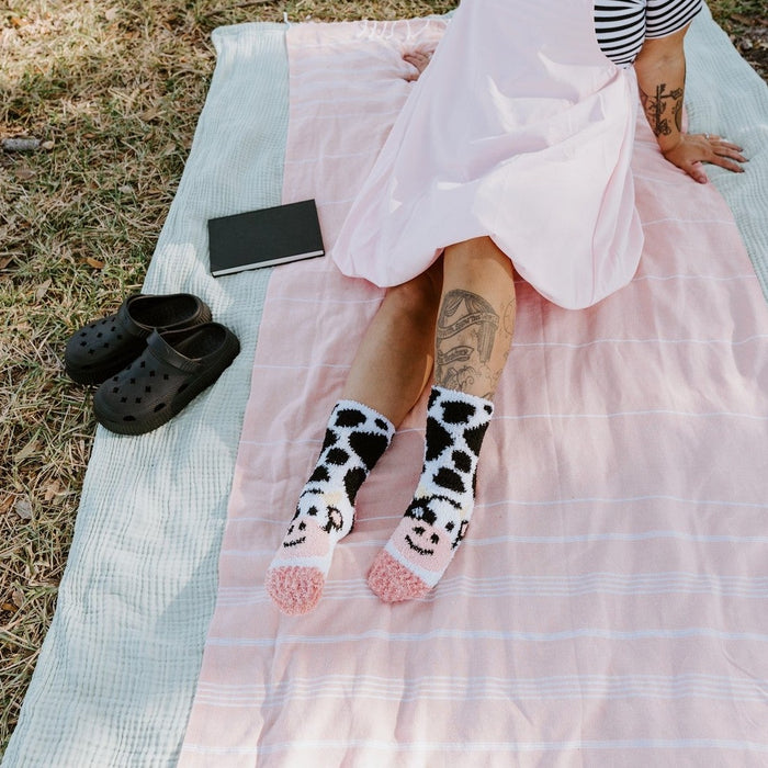 A model leans back on a pink picnic blanket with a soft pink pinafore dress, her Moo Cow Fuzzy socks in White stretched out in front beside a tablet and black Crocs. [lf]
