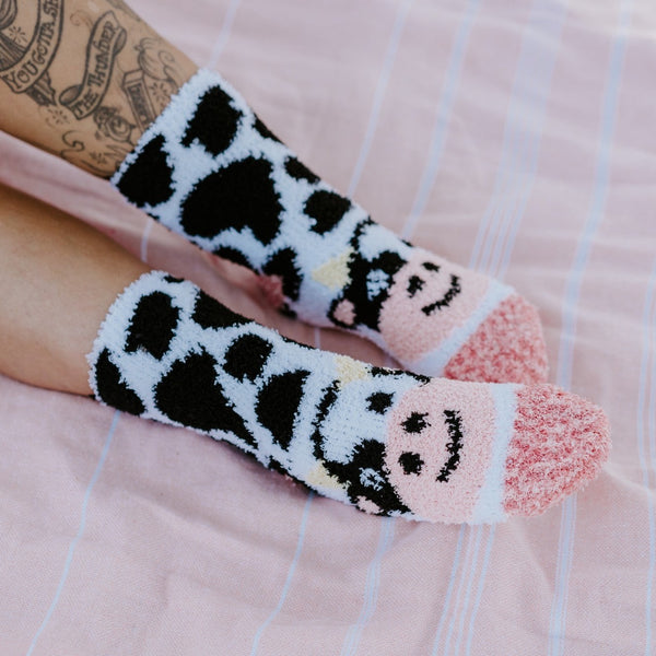 A sunlit close up of tattooed legs and Moo Cow Fuzzy socks in White against a striped pink towel, highlighting the fuzzy cow print and pink smiley toe detail. [lf]