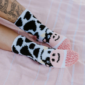 A sunlit close up of tattooed legs and Moo Cow Fuzzy socks in White against a striped pink towel, highlighting the fuzzy cow print and pink smiley toe detail. [lf]