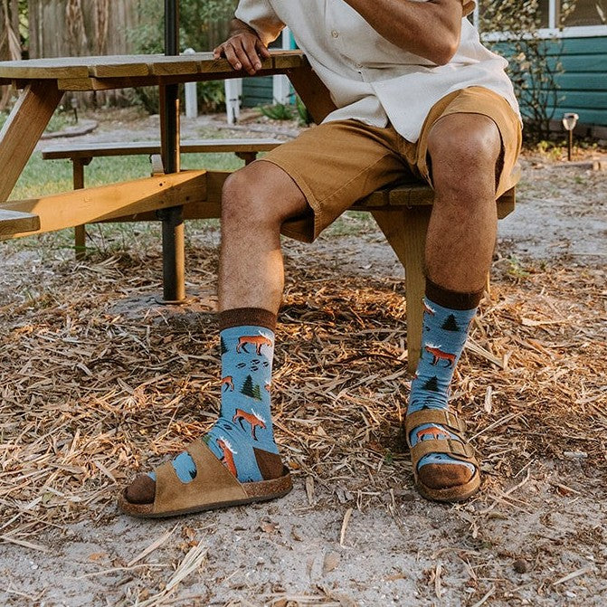 A man in a cream polo and khaki shorts sits at a wooden picnic table under a black patio umbrella, sipping a drink with Moose Tracks Bamboo socks in Blue Heather and brown sandals visible. [lf]