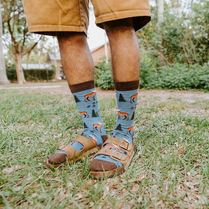 Standing in a backyard with green hedges behind, the model shows the full pattern of Moose Tracks Bamboo socks in Blue Heather tucked into worn brown buckle sandals. [lf]