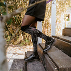 A close side view of a model ascending wooden porch steps in a black dress and black combat boots, with Spider Webs socks in Black pulled to the knee revealing white web patterns. [lf]