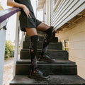 A model leans on a wooden railing and props one foot up on a step, displaying Spider Webs socks in Black with red spider accents above black laced boots in golden light. [lf]