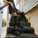 A model leans on a wooden railing and props one foot up on a step, displaying Spider Webs socks in Black with red spider accents above black laced boots in golden light. [lf]