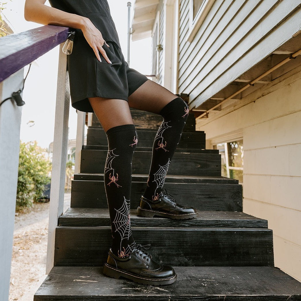 A model leans on a wooden railing and props one foot up on a step, displaying Spider Webs socks in Black with red spider accents above black laced boots in golden light. [lf]