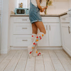 Glancing back over her shoulder with a wide smile, a woman in a denim overall short-set poses beside a sunny kitchen counter holding a fresh bouquet, knee-high Where The Wildflowers Grow socks in Ivory Heather catching the light against pale wood floors. [lf]
