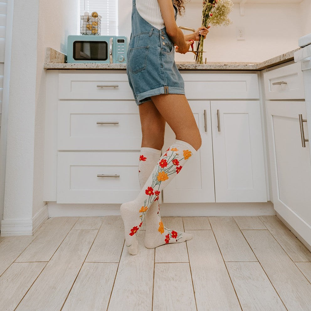 Glancing back over her shoulder with a wide smile, a woman in a denim overall short-set poses beside a sunny kitchen counter holding a fresh bouquet, knee-high Where The Wildflowers Grow socks in Ivory Heather catching the light against pale wood floors. [lf]