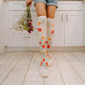 Back-facing crop on light plank flooring frames two legs in knee-high Where The Wildflowers Grow socks in Ivory Heather, one hand at her side trailing a loose bunch of dried wildflowers below cuffed denim shorts. [lf]