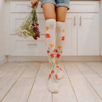Back-facing crop on light plank flooring frames two legs in knee-high Where The Wildflowers Grow socks in Ivory Heather, one hand at her side trailing a loose bunch of dried wildflowers below cuffed denim shorts. [lf]