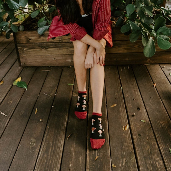 A smiling woman in a red striped blazer sits barefoot-but-socked on a wooden planter ledge surrounded by tropical greenery, showing the Digging the Mushrooms no-show socks in black with red toe and heel accents and tiny toadstool prints. [lf]