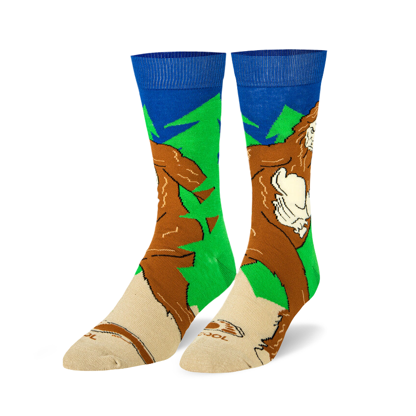 Pair of crew socks featuring a Sasquatch design.  Colors include brown, green, and blue. The Bigfoot image is walking through a forest.