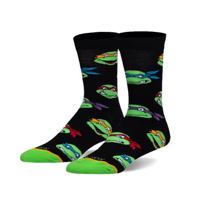 A pair of black crew socks with a repeating pattern of green turtle heads wearing red, blue, orange, and purple masks. Green toe and heel.