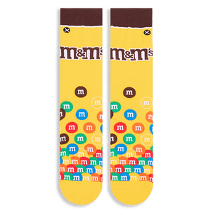 Flat lay of yellow crew socks showing a brown cuff, the M&M's logo on the calf, and colorful M&M candies piled at the toe.