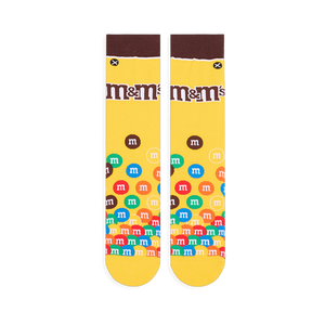 Flat lay of yellow crew socks showing a brown cuff, the M&M's logo on the calf, and colorful M&M candies piled at the toe.
