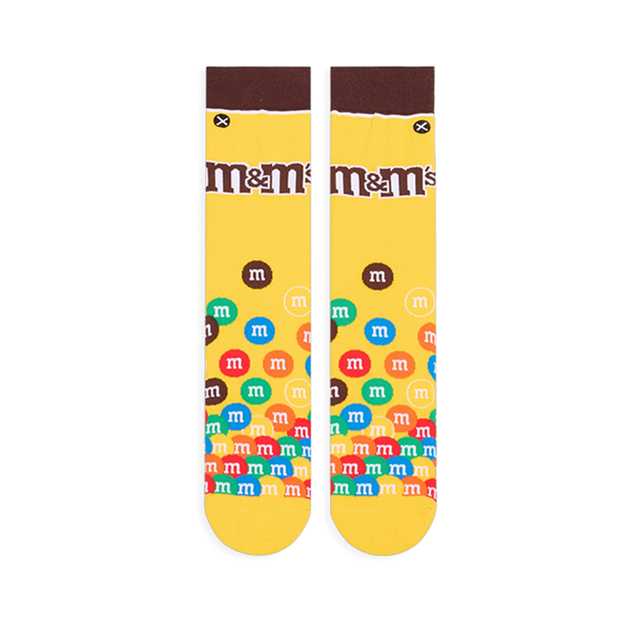 Flat lay of yellow crew socks showing a brown cuff, the M&M's logo on the calf, and colorful M&M candies piled at the toe.