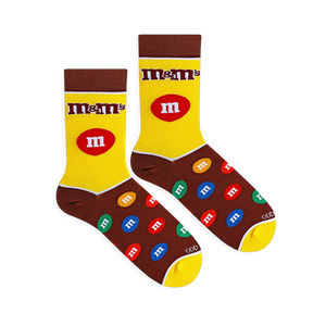 Flat lay of yellow and brown M&M's socks showing the classic logo, a large red candy on the leg, and a colorful candy foot.