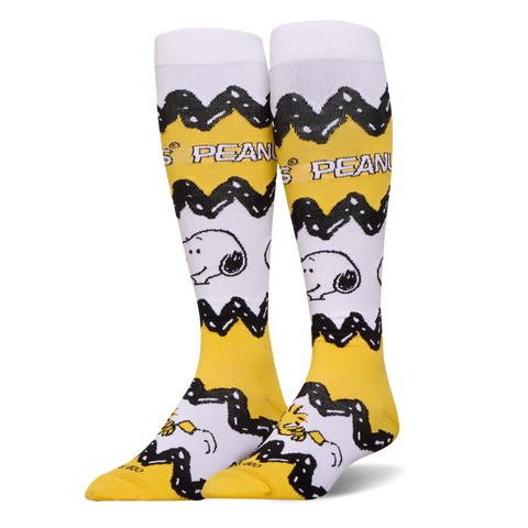Tall yellow and white Peanuts socks featuring Charlie Brown's black zigzag pattern, Snoopy's face, and Woodstock at the toe.