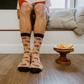 Front view of a person seated on a cushioned window bench reading a book, their Capybara Orange socks in Hemp Heather paired with bare feet on a wood plank floor beside a small pedestal plate of orange slices. [lf]