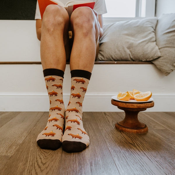 Front view of a person seated on a cushioned window bench reading a book, their Capybara Orange socks in Hemp Heather paired with bare feet on a wood plank floor beside a small pedestal plate of orange slices. [lf]