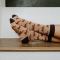 Side profile of bare ankles crossed on a wooden ledge in front of a black wall, the Capybara Orange socks in Hemp Heather displayed to show the full capybara repeat pattern. [lf]