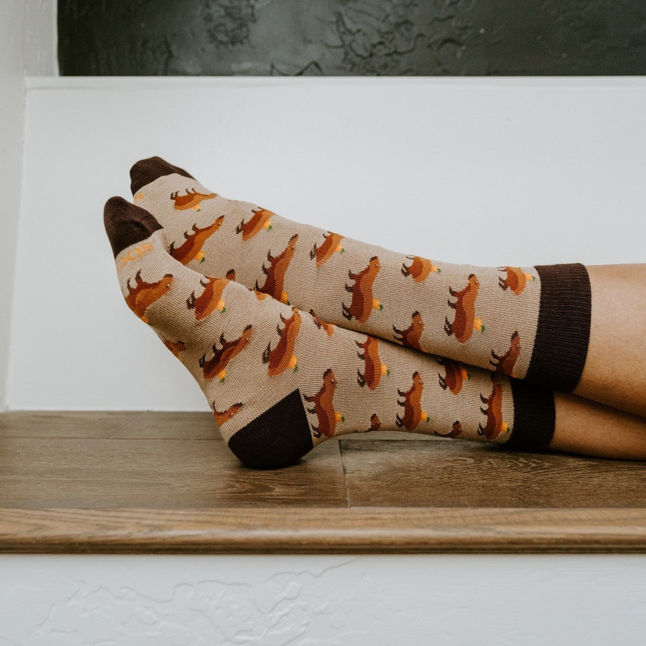 Side profile of bare ankles crossed on a wooden ledge in front of a black wall, the Capybara Orange socks in Hemp Heather displayed to show the full capybara repeat pattern. [lf]