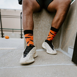 Tight vertical detail of the bat print sock pattern shown from the calves down, paired with cream chunky sneakers on a concrete pavement, wearing Going Batty For Candy socks in Orange. [lf]