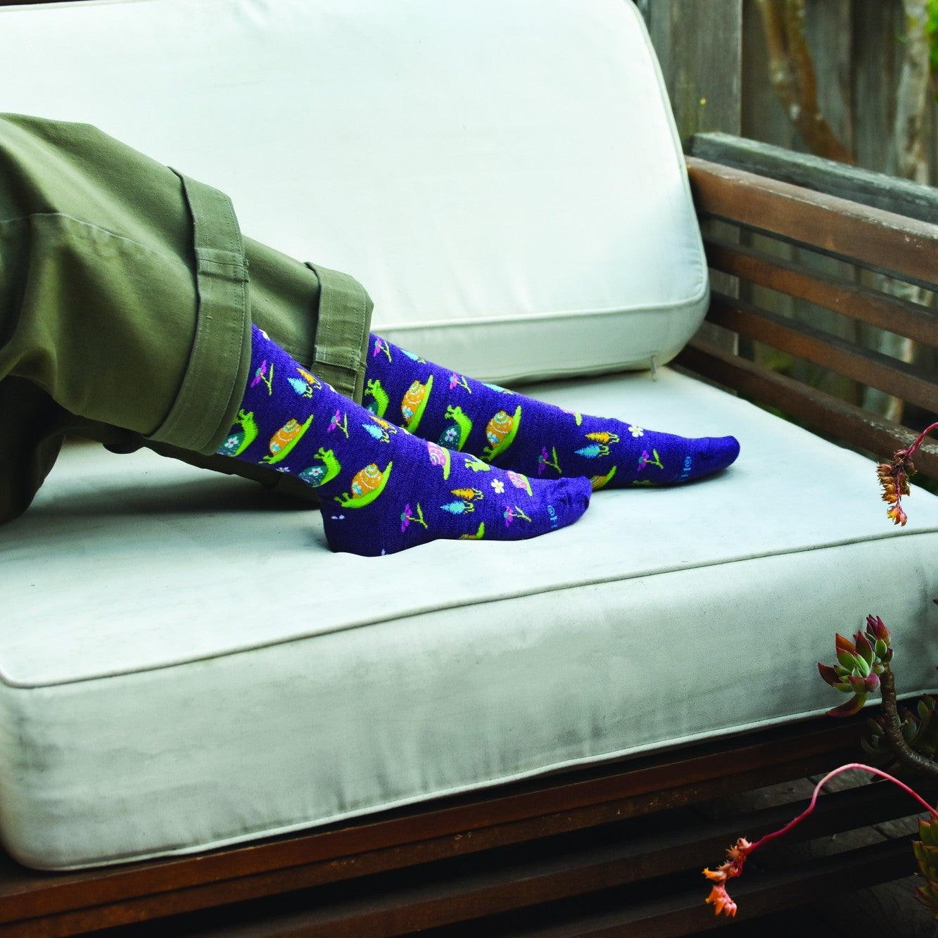 Crossed feet rest on a cream cushioned patio bench next to potted succulents, modeling purple Scenic Snails in Purple Heather socks scattered with colorful snail prints. [lf]