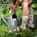 A woman in an olive cardigan crouches in a sunlit garden to lift a galvanized watering can, wearing the Wild Flowers Bamboo socks in Ivory dotted with orange poppies and green leaves. [lf]