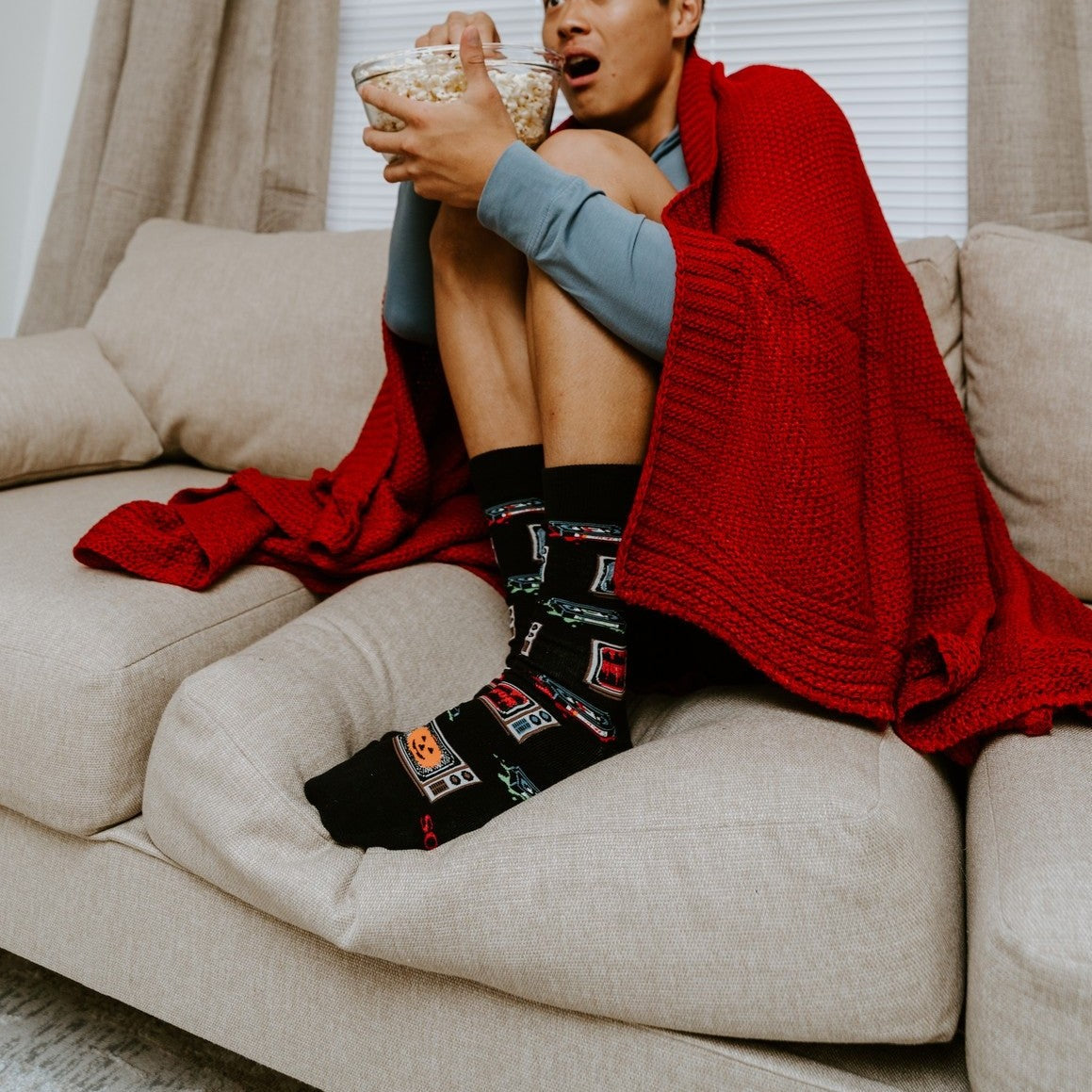 A startled man hugs his knees on the couch with a popcorn bowl raised, wearing 31 Days of Halloween socks in Black featuring pumpkin and creature TV motifs. [lf]