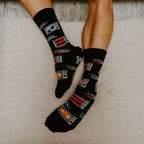 Top-down look at relaxed feet on an ottoman cushion, highlighting the all-over pumpkin and monster pattern of 31 Days of Halloween socks in Black. [lf]