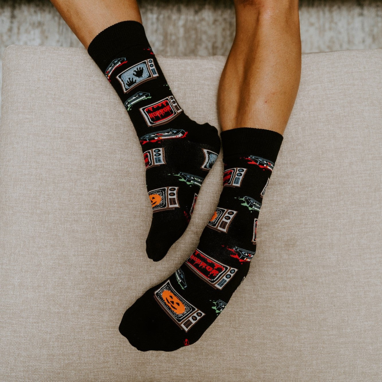 Top-down look at relaxed feet on an ottoman cushion, highlighting the all-over pumpkin and monster pattern of 31 Days of Halloween socks in Black. [lf]