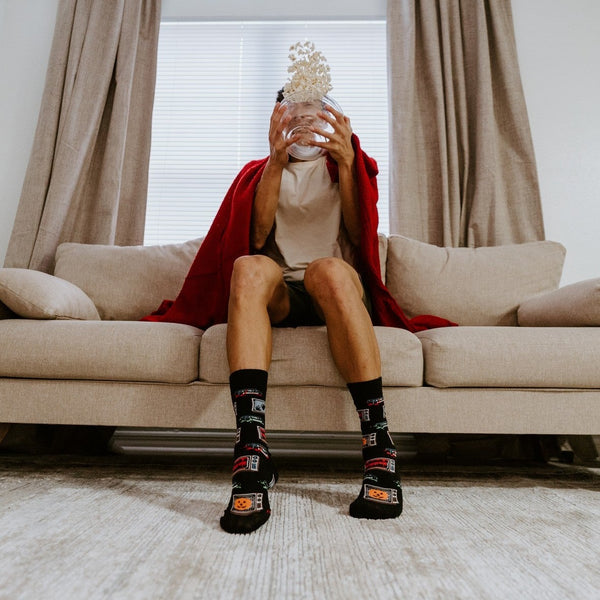 Popcorn tumbles from a glass bowl held up to a man's face on the couch, his planted feet wearing 31 Days of Halloween socks in Black. [lf]