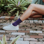 A woman in a white crop top, denim shorts and sage jacket sits on a brick step among tropical plants, her purple Solid Bamboo socks and white sneakers crossed in front. [lf]