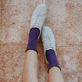 Overhead shot of a woman's bare legs raised against a tan tile wall, her white sneakers and purple Solid Bamboo socks pointed toward the ceiling. [lf]