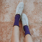 Overhead shot of a woman's bare legs raised against a tan tile wall, her white sneakers and purple Solid Bamboo socks pointed toward the ceiling. [lf]