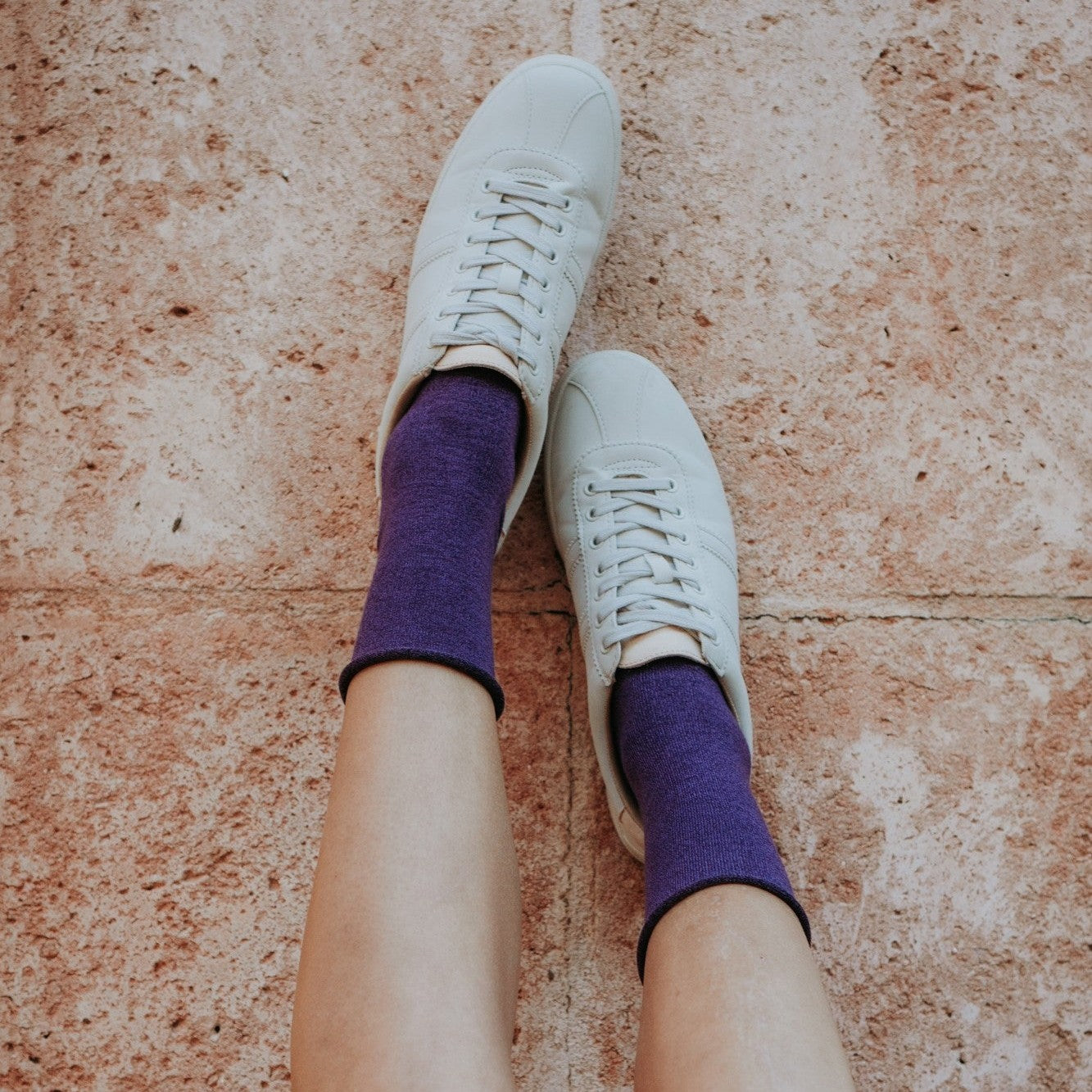 Overhead shot of a woman's bare legs raised against a tan tile wall, her white sneakers and purple Solid Bamboo socks pointed toward the ceiling. [lf]