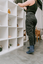 Side view of a woman in dark jeans and a black tank top arranging items on a tall white shelving unit, her navy Solid Bamboo socks peeking out beneath the cuffs. [lf]