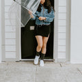 A woman in a denim jacket and black mini skirt stands in a doorway holding a clear bubble umbrella, her black Solid Bamboo socks pulled up to mid-calf above white sneakers. [lf]