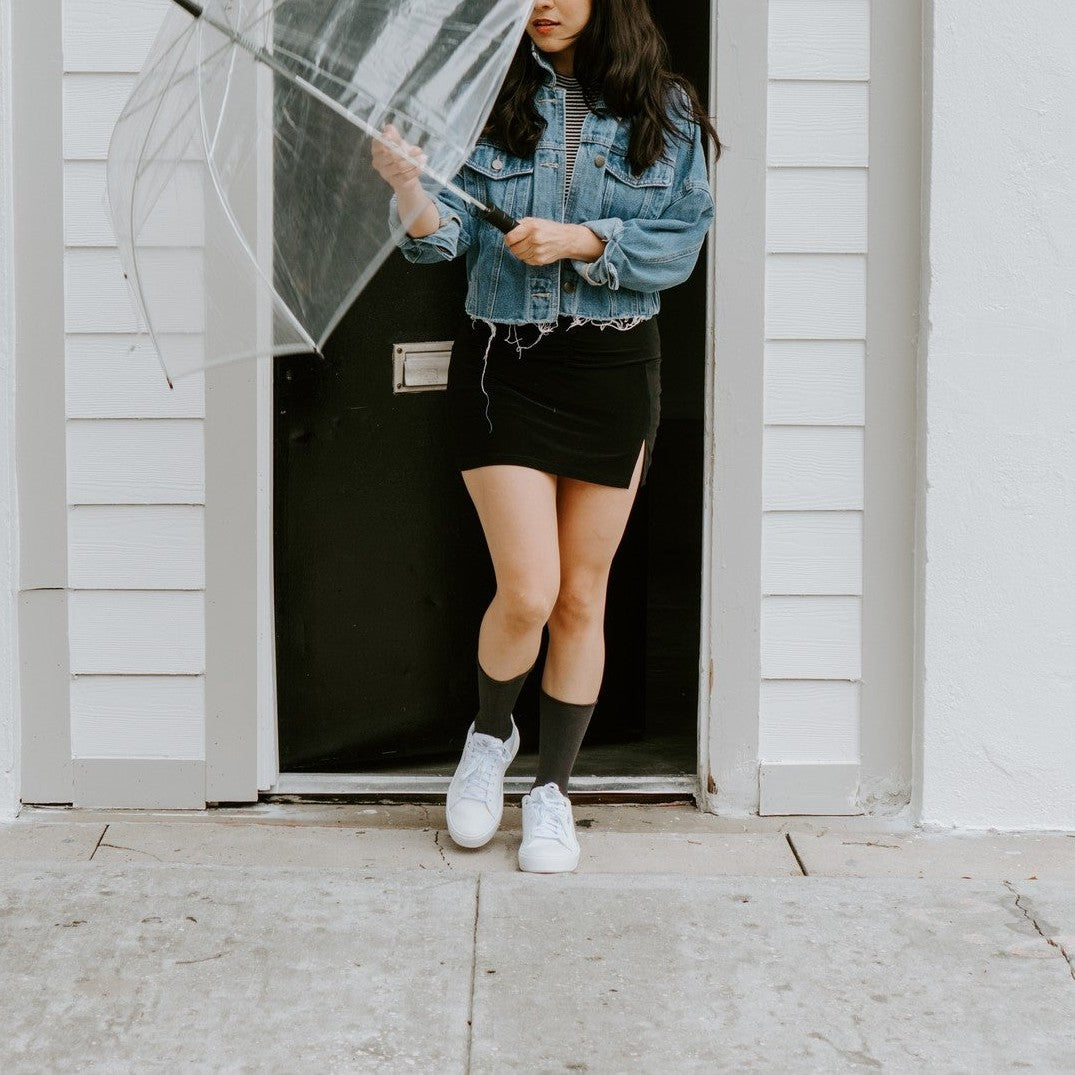 A woman in a denim jacket and black mini skirt stands in a doorway holding a clear bubble umbrella, her black Solid Bamboo socks pulled up to mid-calf above white sneakers. [lf]