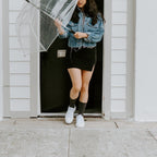 A woman in a denim jacket and black mini skirt stands in a doorway holding a clear bubble umbrella, her black Solid Bamboo socks pulled up to mid-calf above white sneakers. [lf]