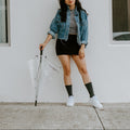 A woman in a cropped denim jacket and black skirt poses against a white wall holding a clear umbrella by its handle, her black Solid Bamboo socks crossed above white sneakers. [lf]