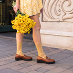 A woman in a yellow floral mini dress carries a bouquet of yellow chrysanthemums past a stone storefront wearing Solid Bamboo socks in honey gold and tan leather loafers. [lf]