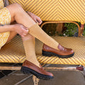 A woman in a yellow floral dress sits on a woven bistro bench adjusting her Solid Bamboo socks in honey gold, paired with brown leather lug-sole loafers. [lf]