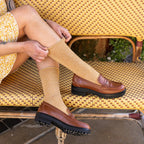 A woman in a yellow floral dress sits on a woven bistro bench adjusting her Solid Bamboo socks in honey gold, paired with brown leather lug-sole loafers. [lf]