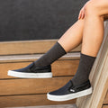Legs crossed over the edge of a wooden bench show Solid Bamboo socks in charcoal grey heather pulled up over black perforated slip-on sneakers against a dark wall. [lf]