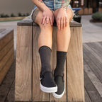 A heavily tattooed woman in a white tee and denim shorts perches on a wooden plaza box wearing Solid Bamboo socks in charcoal grey heather and black slip-on sneakers. [lf]
