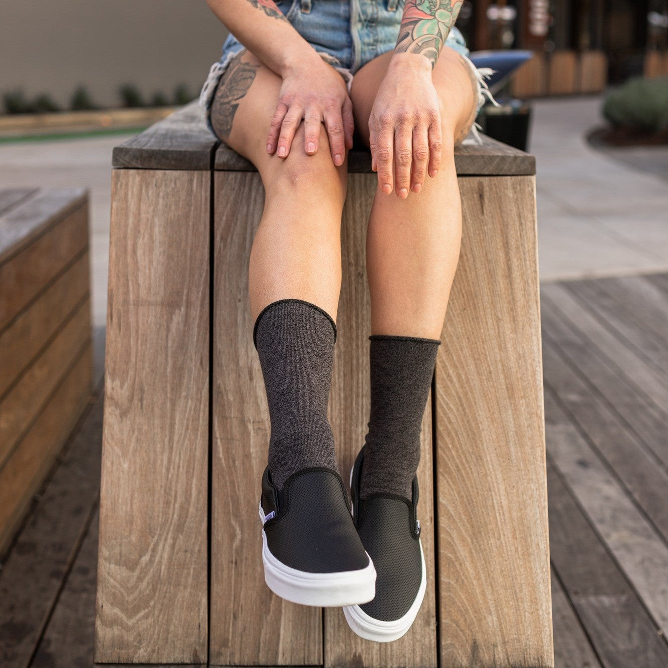 A heavily tattooed woman in a white tee and denim shorts perches on a wooden plaza box wearing Solid Bamboo socks in charcoal grey heather and black slip-on sneakers. [lf]
